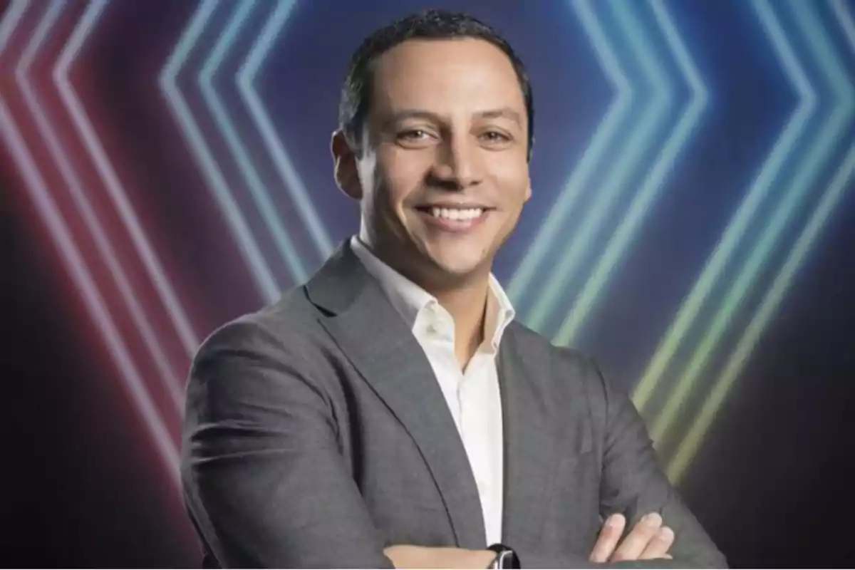 Hombre de cabello corto y oscuro con saco gris y camisa blanca sonriendo frente a un fondo con luces de neón en forma de flechas Hombre de cabello corto y oscuro con saco gris y camisa blanca sonriendo frente a un fondo con luces de neón en forma de flechas