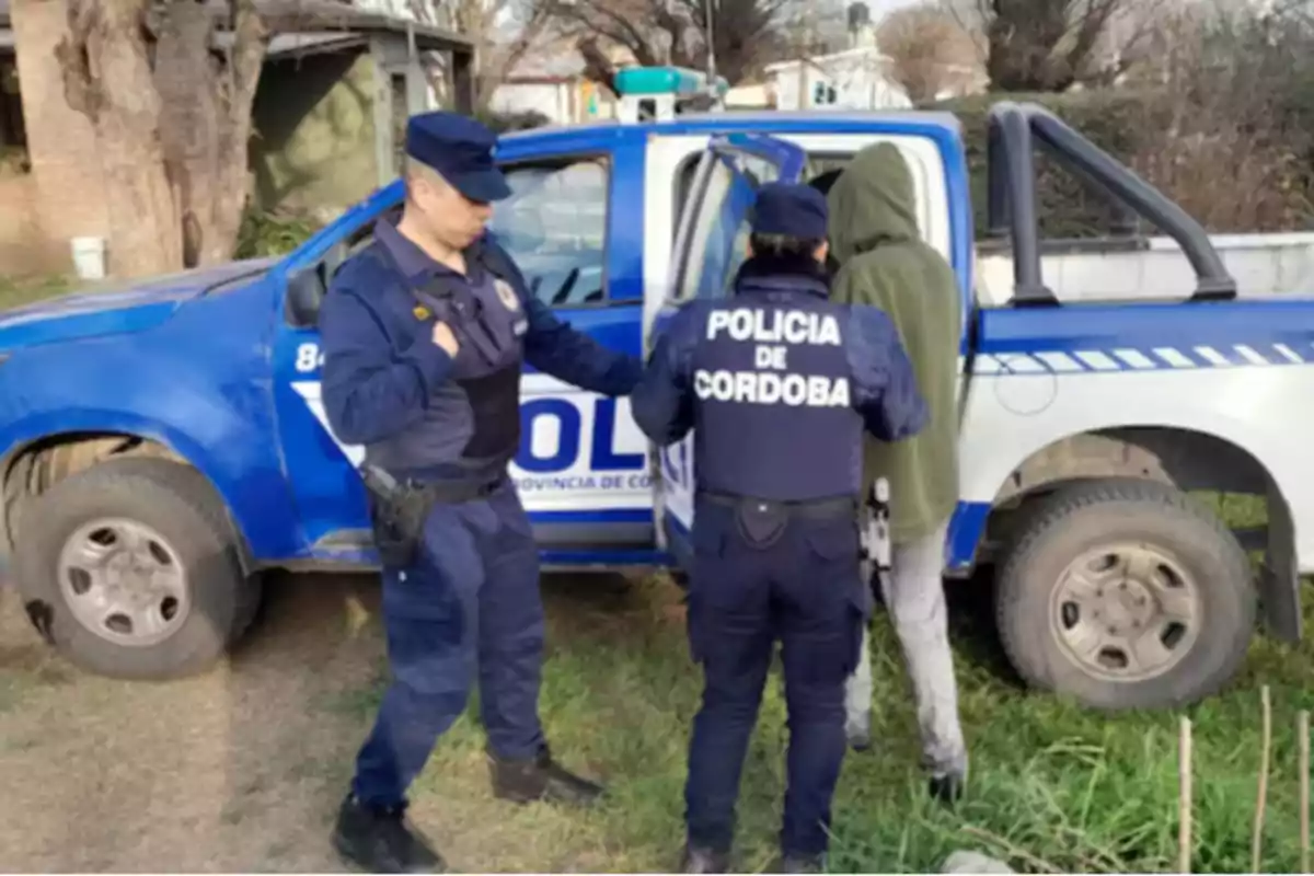 Two Córdoba police officers detain a person wearing a green sweatjersey next to a blue police truck in an outdoor setting.