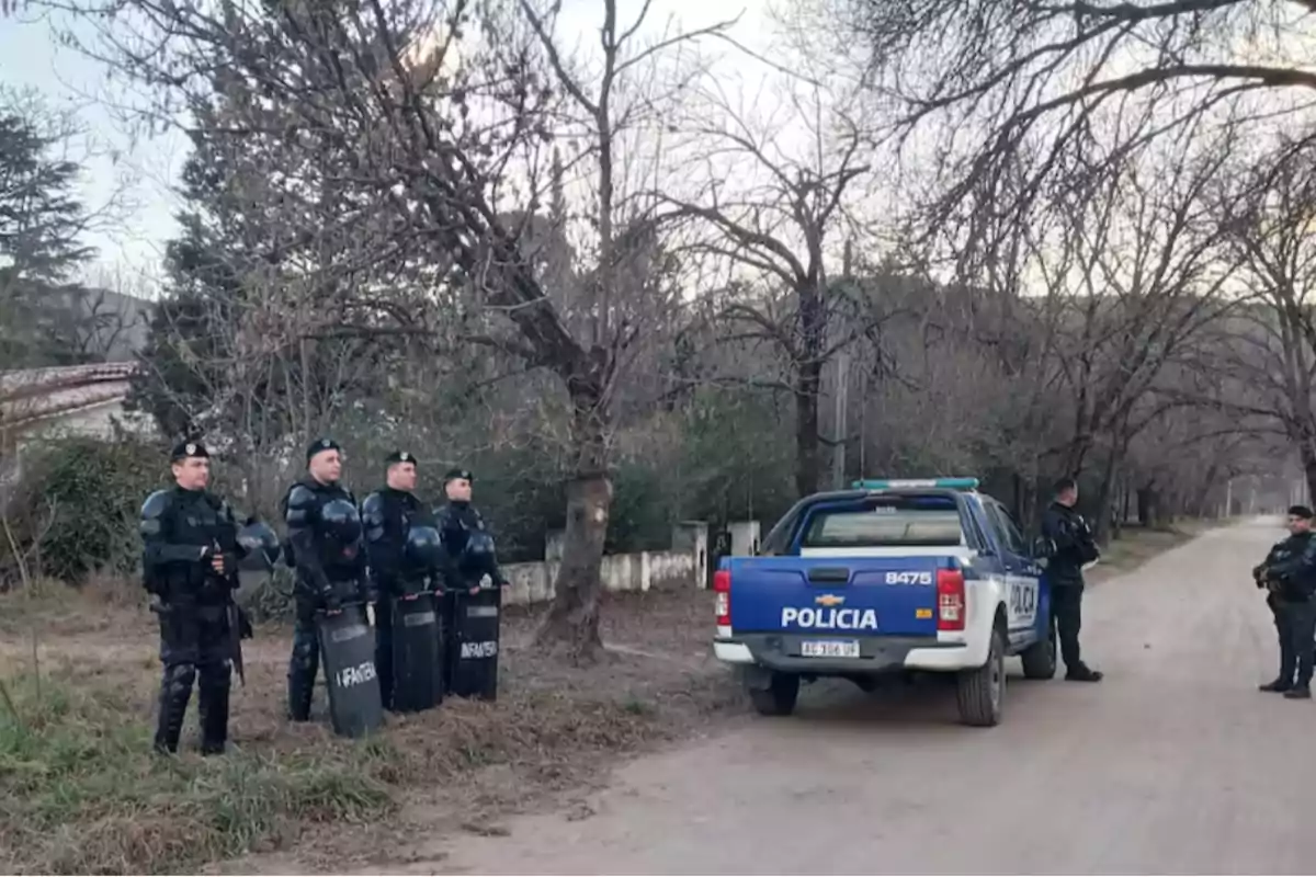 Several police officers in riot gear are standing next to a blue police van on a dirt road surrounded by leafless trees. Several police officers in riot gear are standing next to a blue police van on a dirt road surrounded by leafless trees.