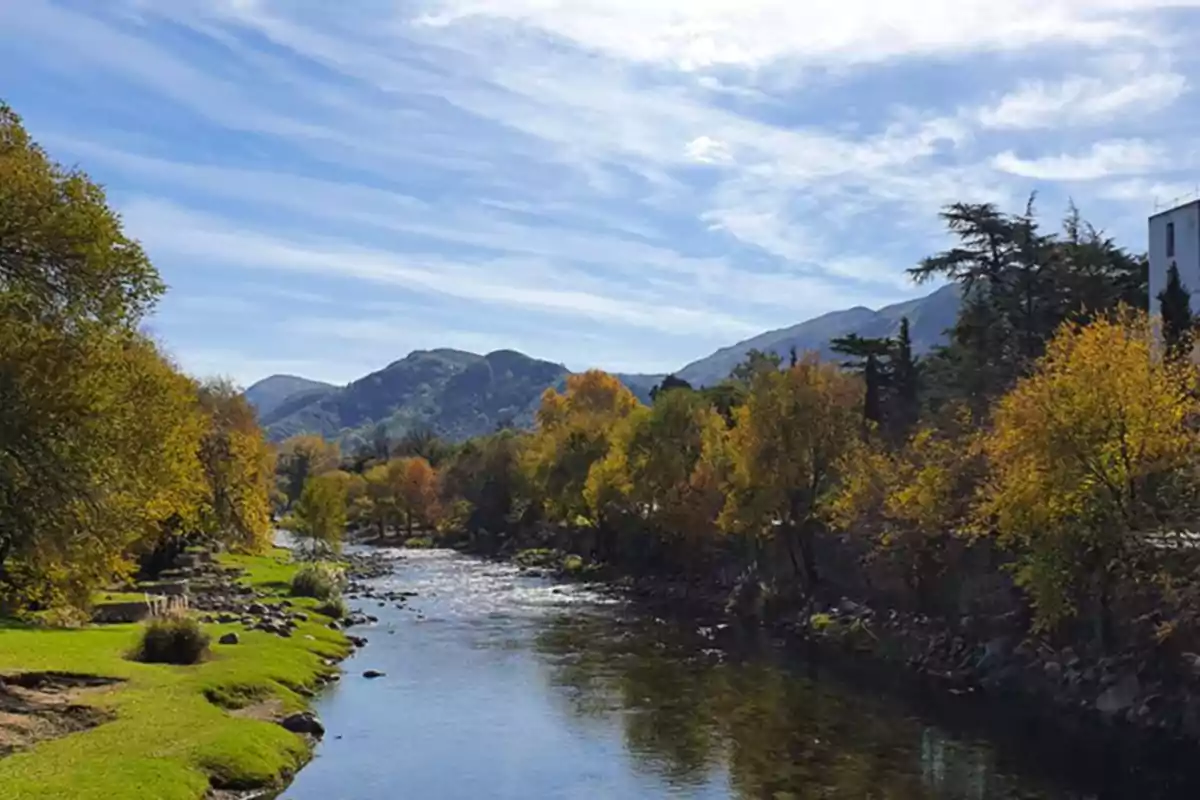 River surrounded by trees with autumn foliage and mountains in the background under a blue sky with scattered clouds River surrounded by trees with autumn foliage and mountains in the background under a blue sky with scattered clouds