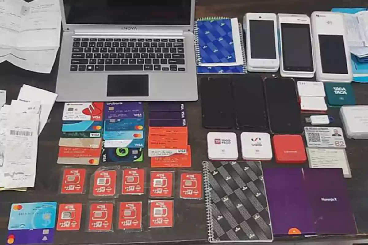 On a table, there is a laptop, several mobile phones, bank cards, SIM cards, receipts, notebooks, and electronic devices arranged in rows.