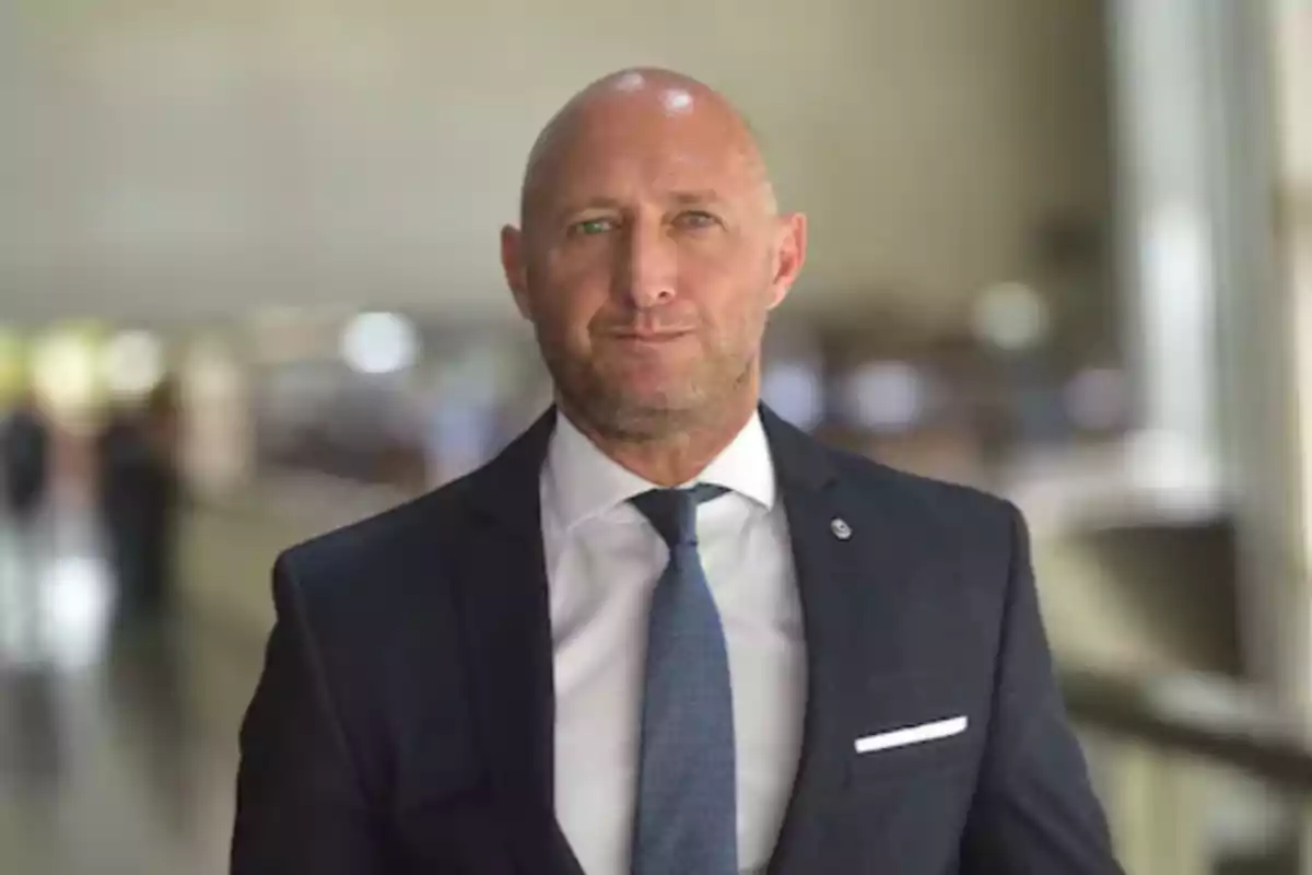 Bald man with a short beard wearing a blue suit and tie, posing in a blurred professional setting. Bald man with a short beard wearing a blue suit and tie, posing in a blurred professional setting.