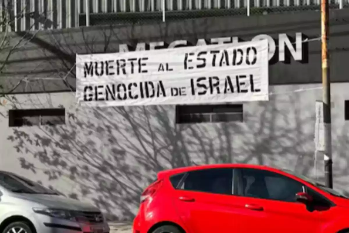 Pancarta con mensaje político en una pared gris sobre unos autos estacionados