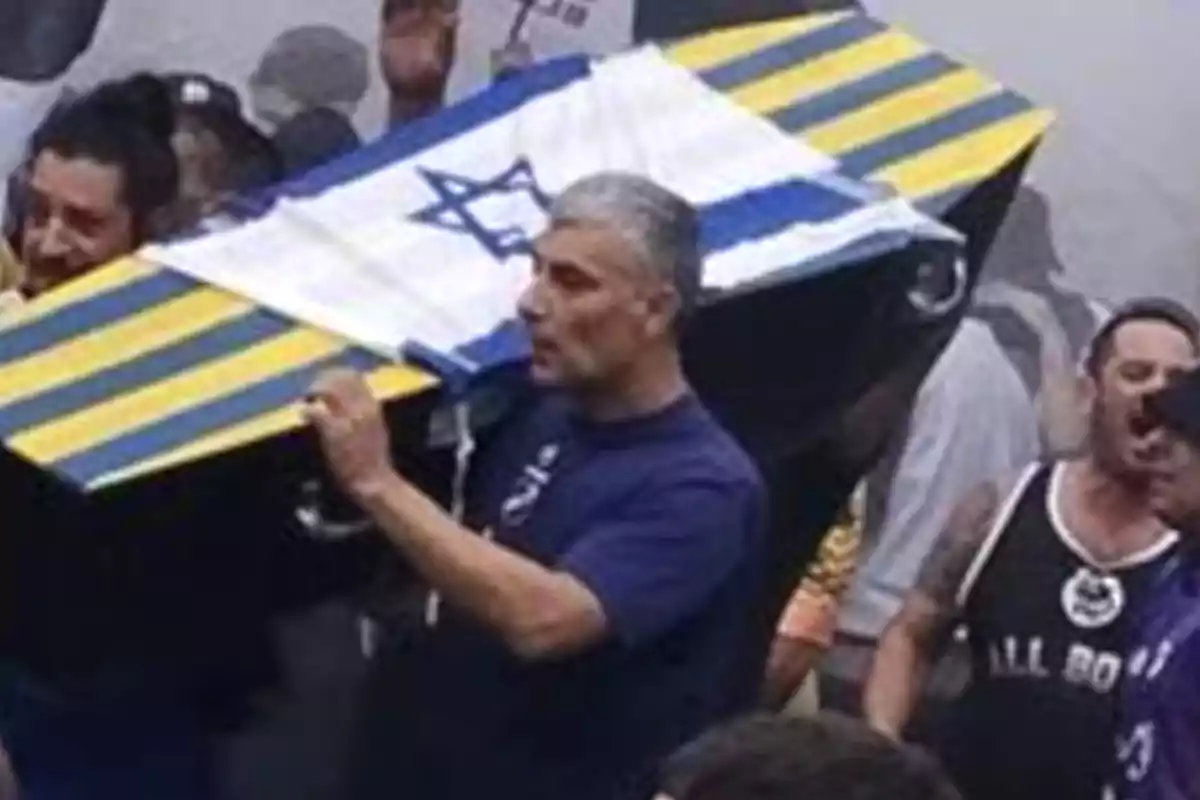 Un grupo de personas lleva un ataúd cubierto con una bandera de Israel y decorado con franjas amarillas y azules en un evento multitudinario. Un grupo de personas lleva un ataúd cubierto con una bandera de Israel y decorado con franjas amarillas y azules en un evento multitudinario.
