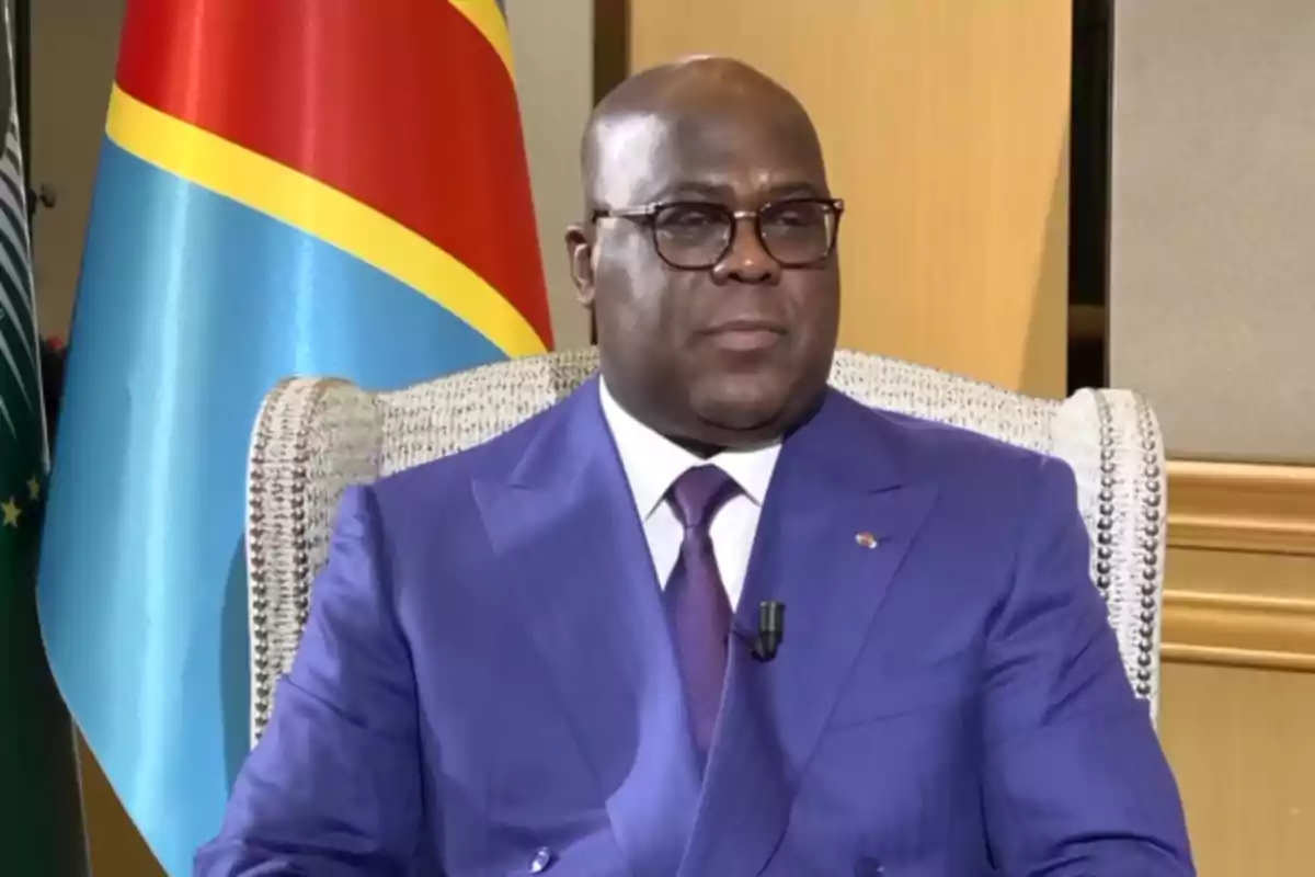 Hombre con gafas y traje azul sentado en una silla frente a una bandera de la República Democrática del Congo Hombre con gafas y traje azul sentado en una silla frente a una bandera de la República Democrática del Congo