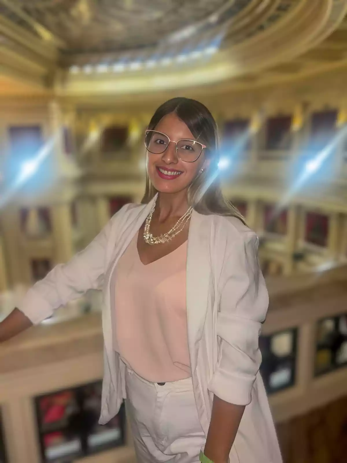 Young woman with glasses and stylish light-colored clothing smiling inside a historic building with classical architecture and bright lights in the background Young woman with glasses and stylish light-colored clothing smiling inside a historic building with classical architecture and bright lights in the background