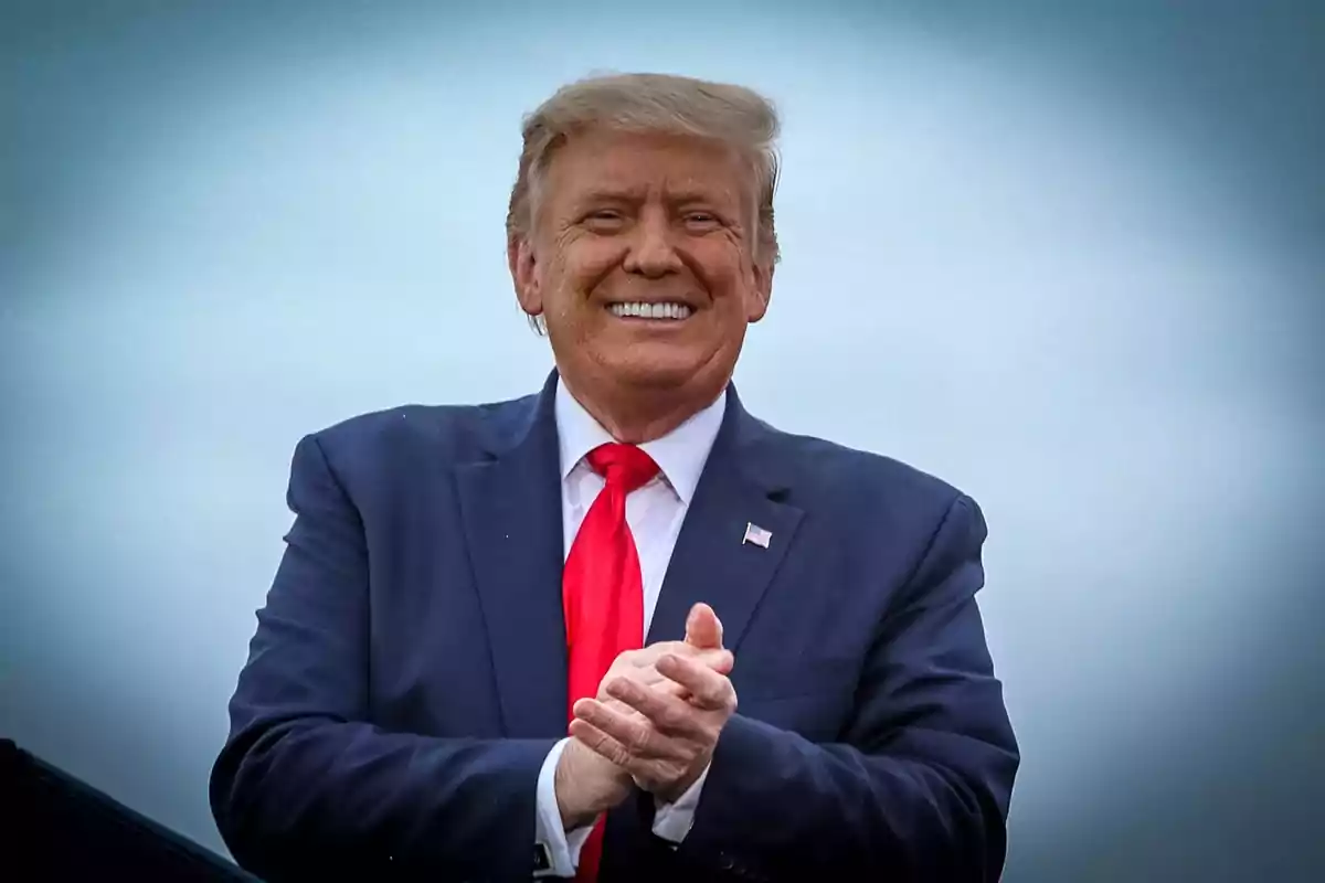 Hombre de cabello rubio con traje azul y corbata roja sonriendo y aplaudiendo frente a un fondo azul claro