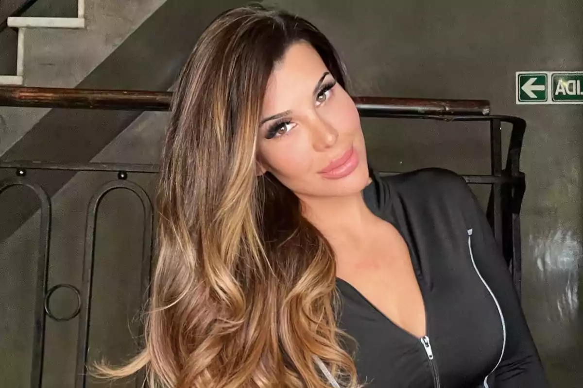 Mujer de cabello largo y ondulado con mechas rubias, vestida con un top negro de cierre, posando y sonriendo frente a una barandilla en un ambiente interior