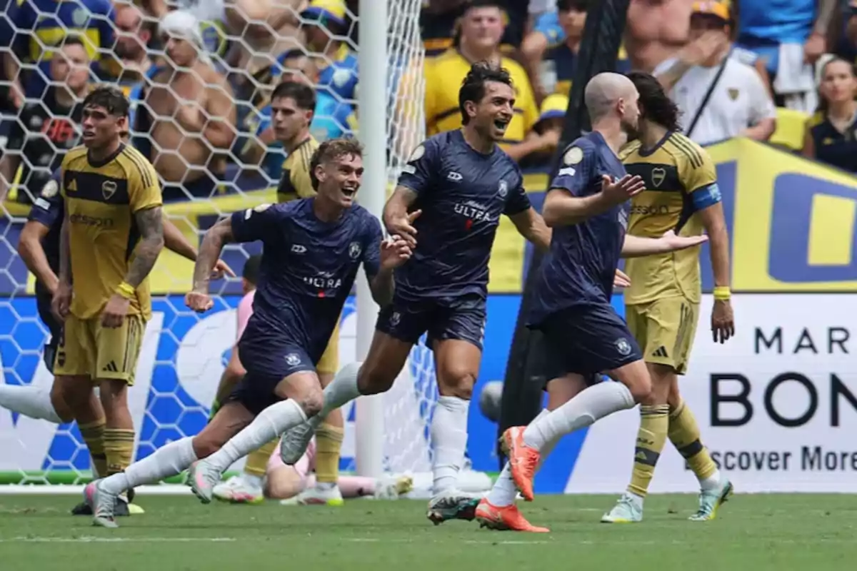 Players from a soccer team celebrate a goal while their opponents show disappointment near the goal during a match in a stadium full of fans.
