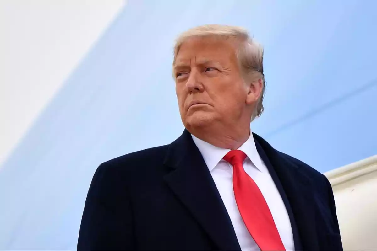 Hombre de cabello rubio con traje oscuro y corbata roja mirando hacia un lado con fondo azul claro Hombre de cabello rubio con traje oscuro y corbata roja mirando hacia un lado con fondo azul claro