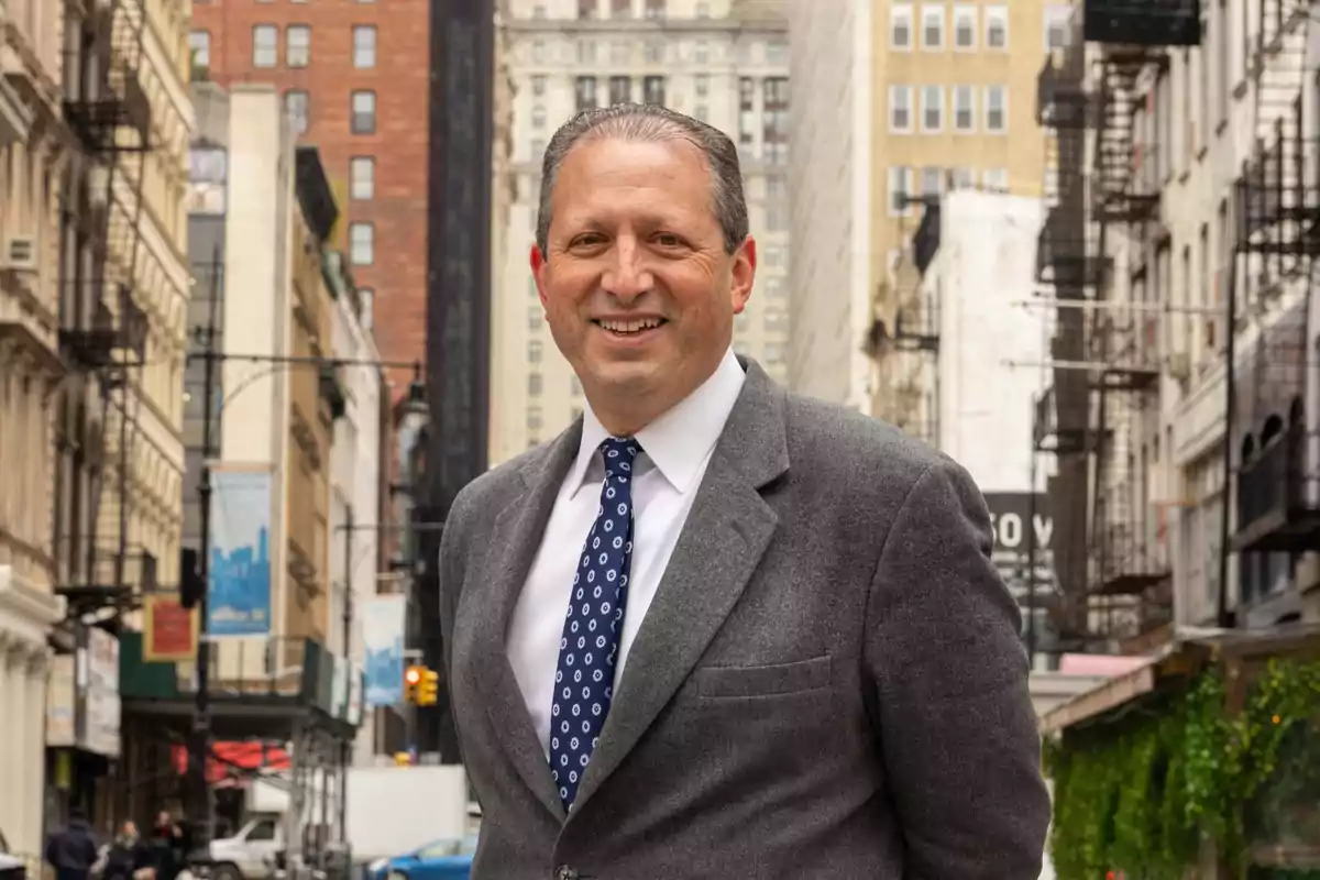 Hombre de mediana edad con traje gris y corbata azul posando y sonriendo en una calle urbana con edificios altos al fondo Hombre de mediana edad con traje gris y corbata azul posando y sonriendo en una calle urbana con edificios altos al fondo
