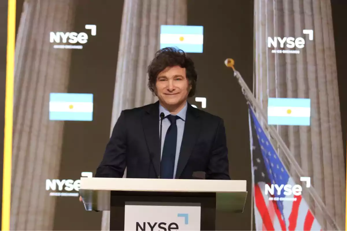 A man with curly hair and a dark suit is standing behind a podium with the NYSE logo; in the background, there are Argentine and U.S. flags next to large columns.