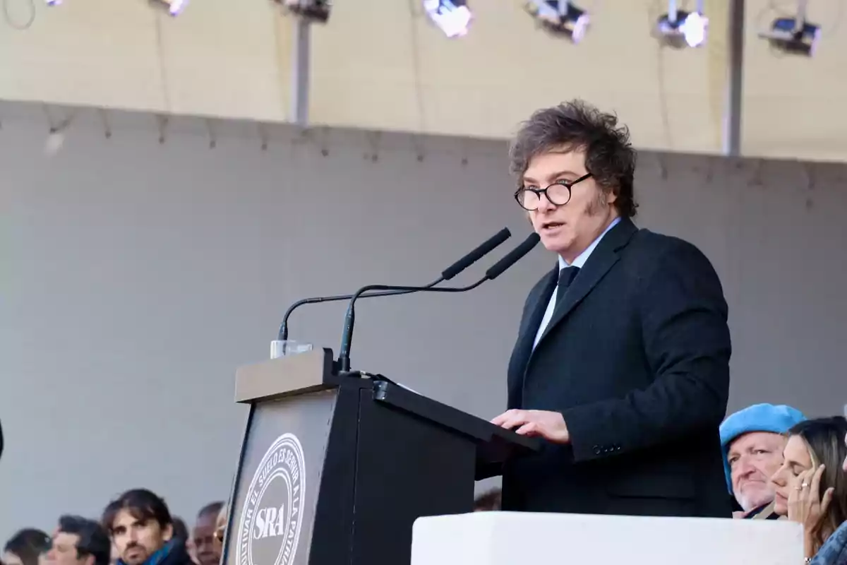 A man wearing glasses and a dark suit speaks at a podium during an outdoor public event while several people watch him from the audience.
