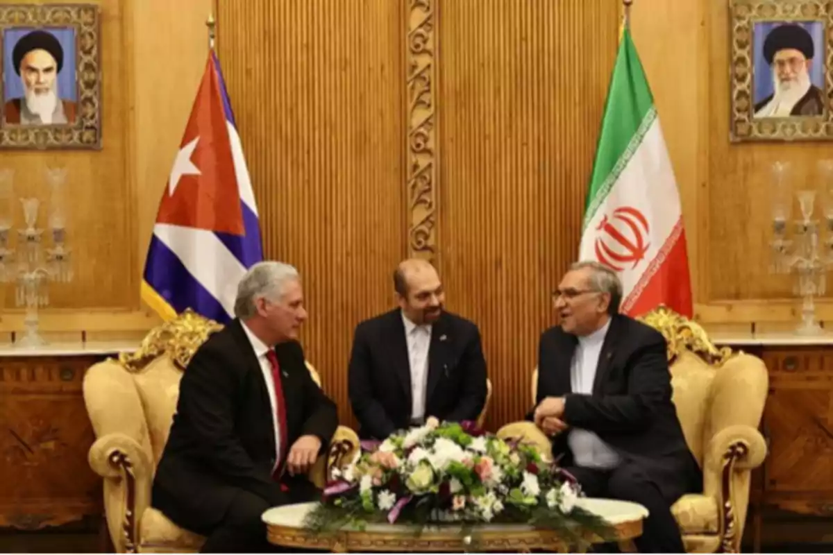 Three men in formal suits are seated and conversing in an elegant room with the flags of Cuba and Iran behind them and a floral arrangement on the table. Three men in formal suits are seated and conversing in an elegant room with the flags of Cuba and Iran behind them and a floral arrangement on the table.
