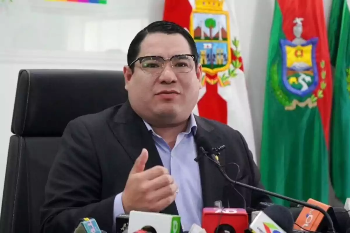 Man with glasses and a dark jacket speaking at a press conference with microphones and official flags in the background Man with glasses and a dark jacket speaking at a press conference with microphones and official flags in the background