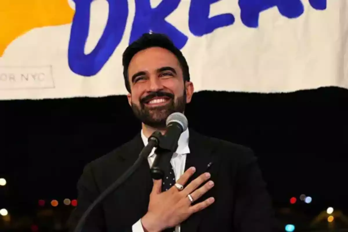 Smiling man with a beard and suit speaking in front of a microphone with one hand on his chest and a sign in the background