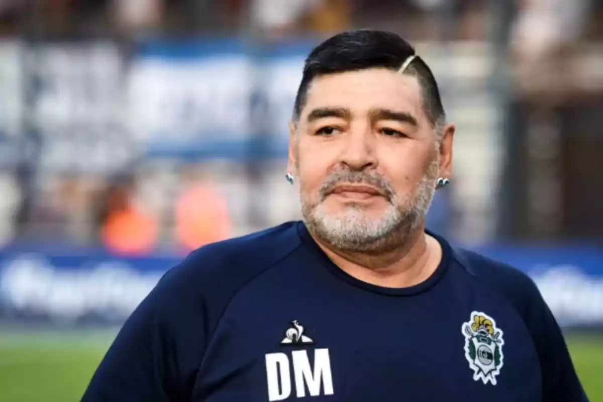 Hombre de cabello corto y barba canosa vistiendo una camiseta azul con las iniciales DM y un escudo deportivo, en un estadio de fútbol.