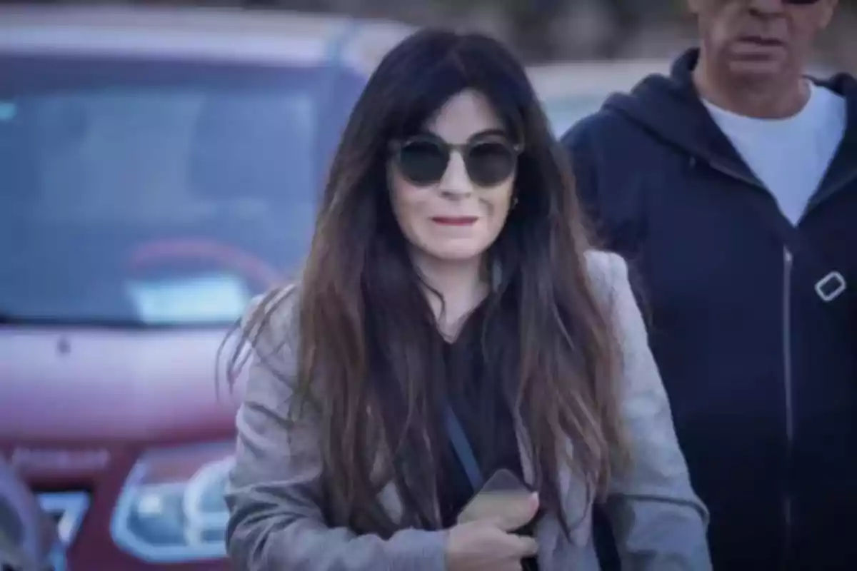 Mujer de cabello largo y oscuro con gafas de sol y chaqueta clara sosteniendo un teléfono móvil frente a un automóvil rojo Mujer de cabello largo y oscuro con gafas de sol y chaqueta clara sosteniendo un teléfono móvil frente a un automóvil rojo