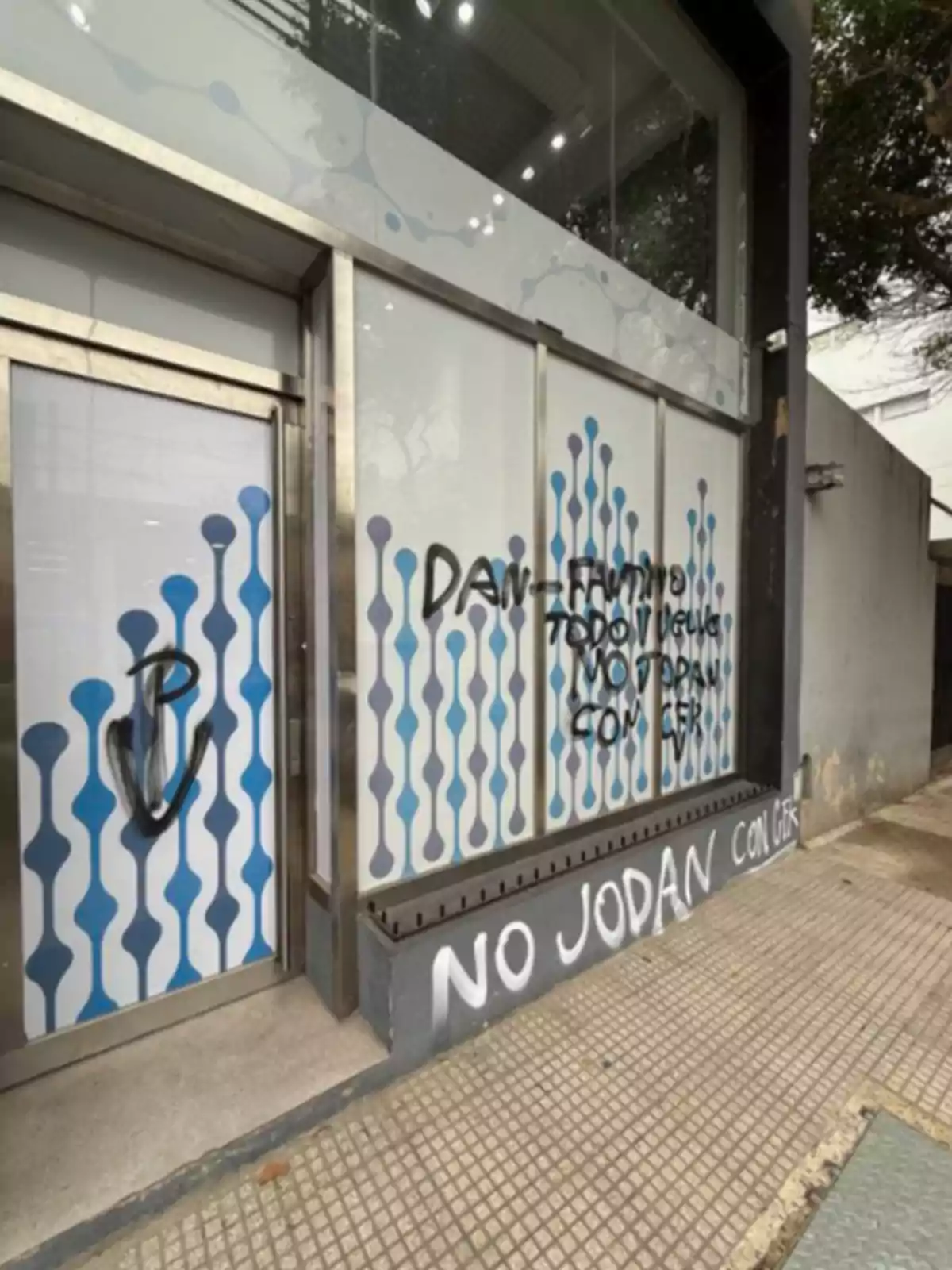 Fachada de un local con vidrios decorados y varios grafitis en aerosol negro y blanco, incluyendo frases de protesta y un dibujo en la puerta Fachada de un local con vidrios decorados y varios grafitis en aerosol negro y blanco, incluyendo frases de protesta y un dibujo en la puerta