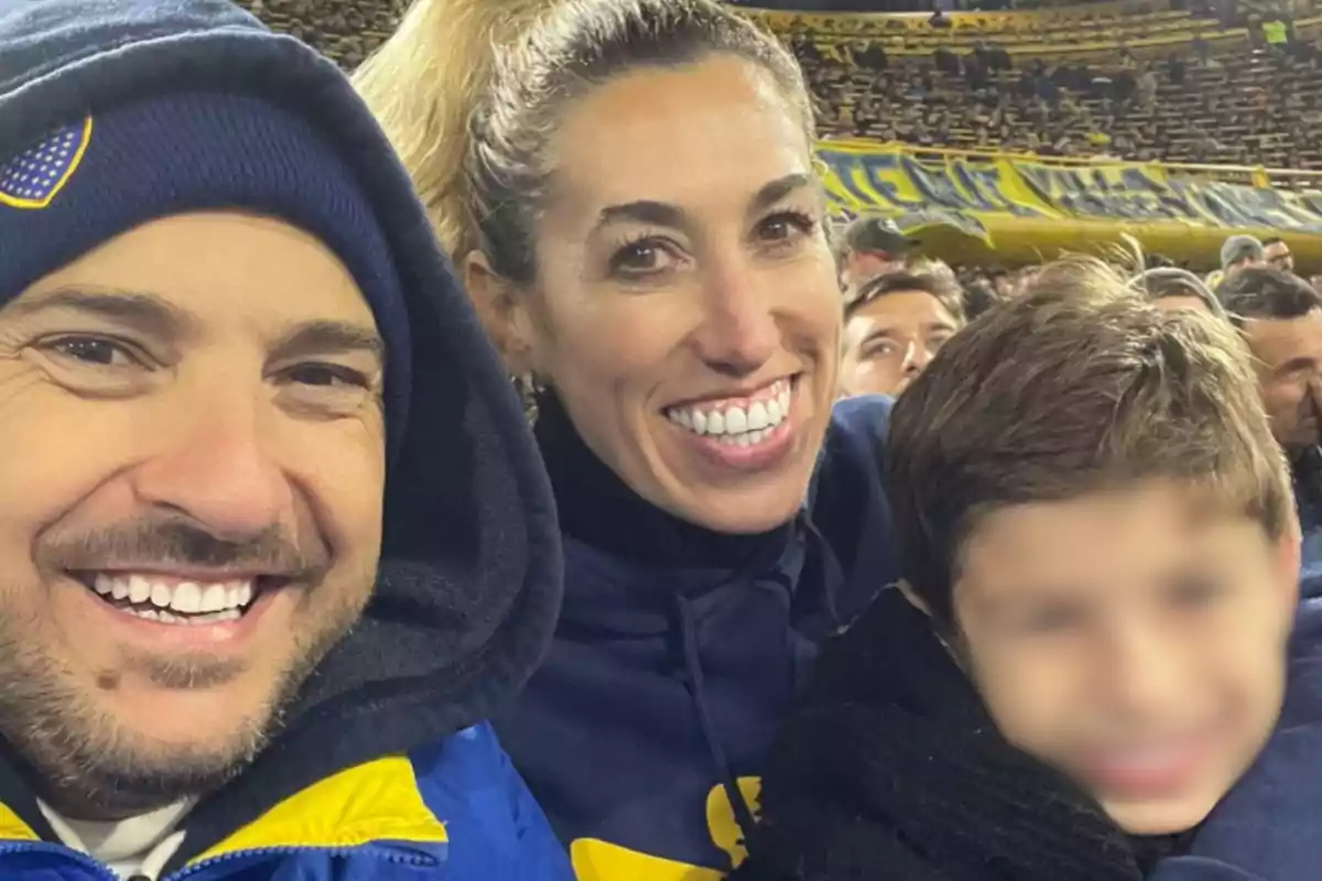 Una familia sonríe y posa para una foto en un estadio de fútbol lleno de gente con banderas amarillas y azules en las gradas. Una familia sonríe y posa para una foto en un estadio de fútbol lleno de gente con banderas amarillas y azules en las gradas.