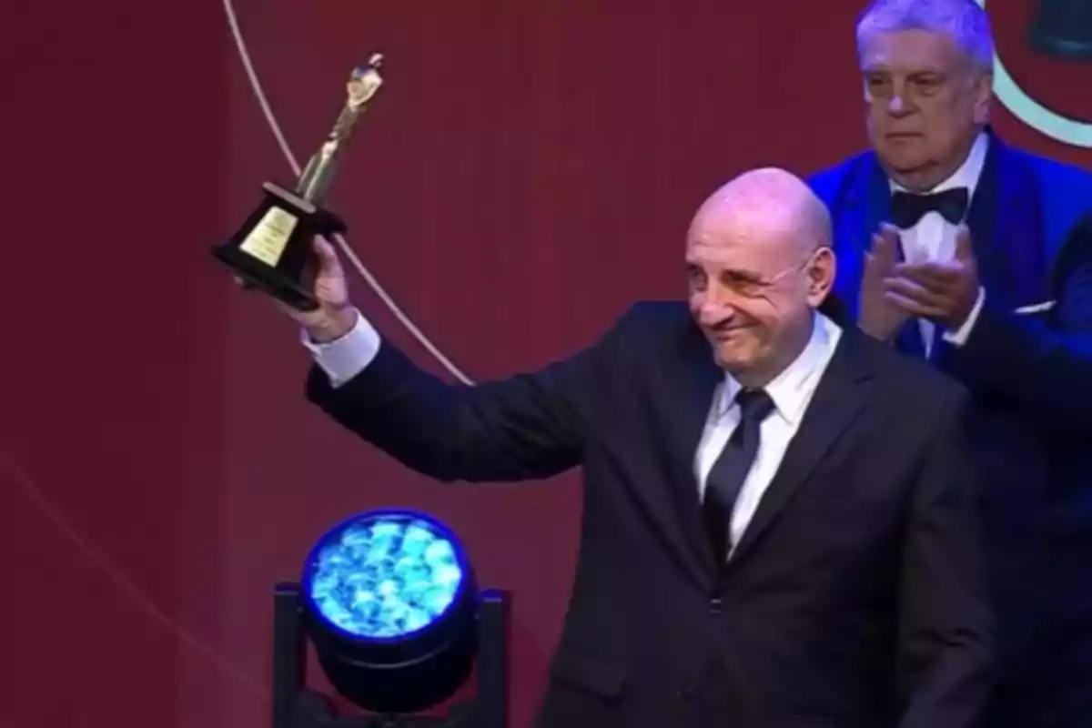 Bald man in a suit smiles and raises a trophy while another person applauds in the background during an award ceremony.