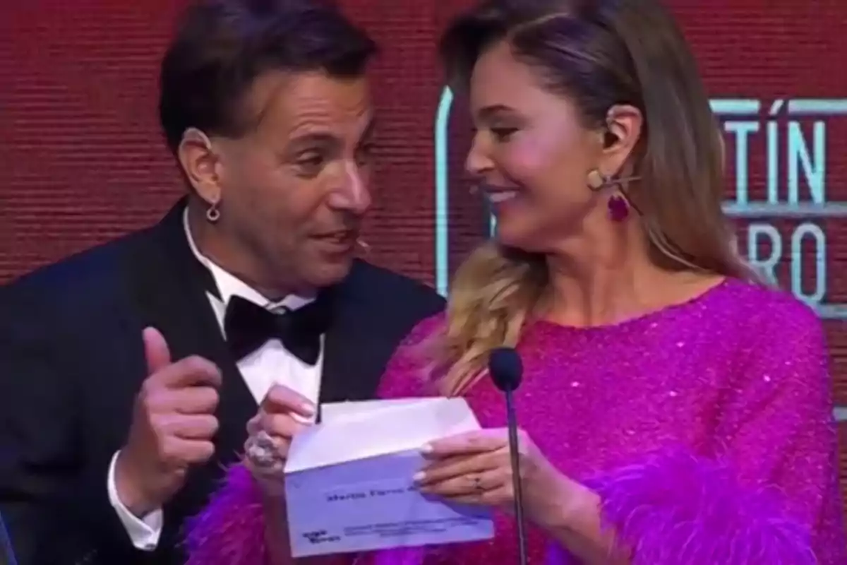 Two elegantly dressed people smile as they announce an award at an event in front of a microphone. Two elegantly dressed people smile as they announce an award at an event in front of a microphone.