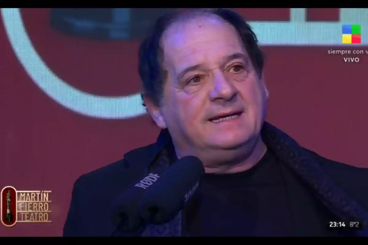 Man speaking in front of a microphone at a theater awards ceremony with a red background and Martín Fierro Teatro logo. Man speaking in front of a microphone at a theater awards ceremony with a red background and Martín Fierro Teatro logo.