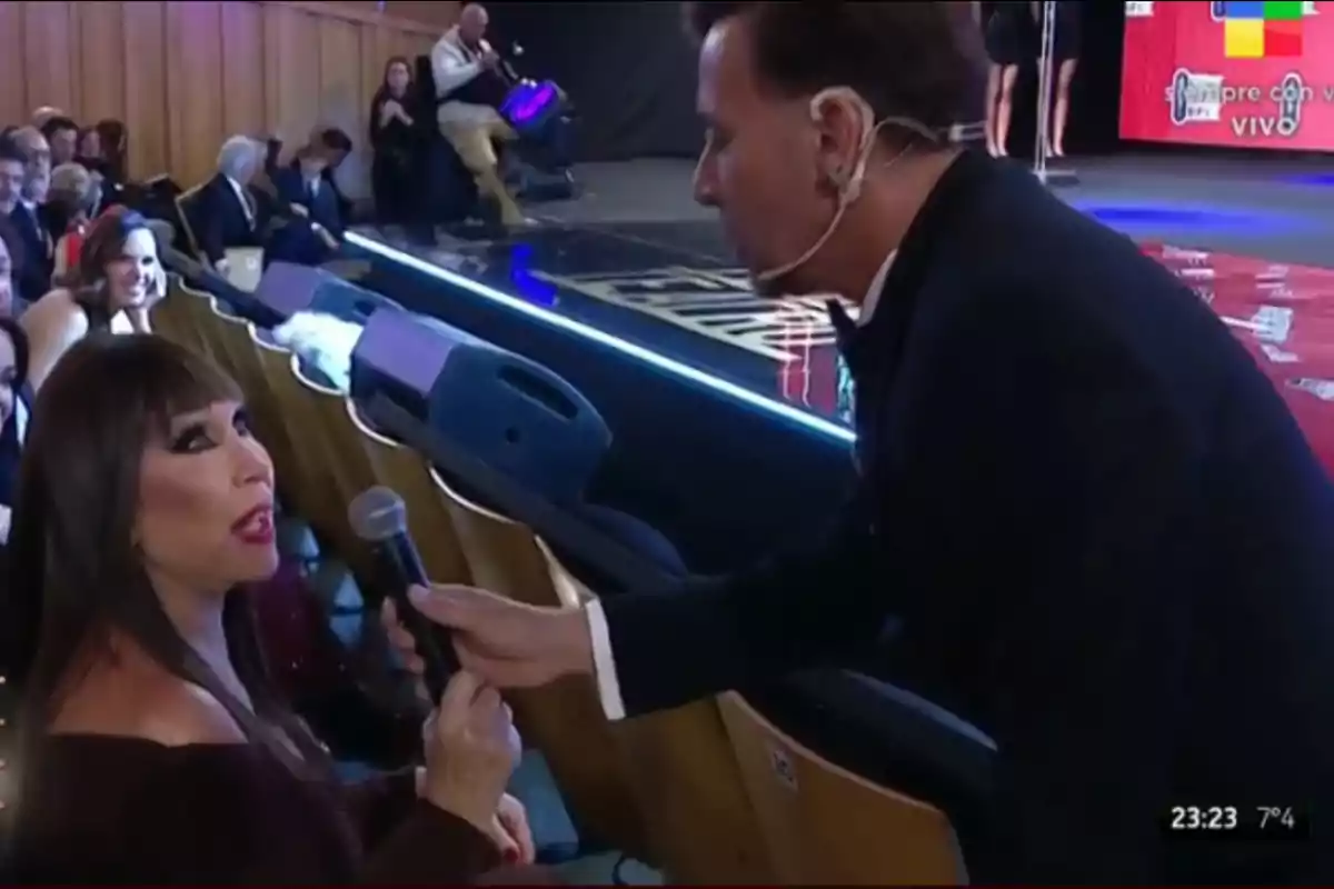 A woman sitting in an armchair holds a microphone while a well-dressed man leans toward her at a live event. A woman sitting in an armchair holds a microphone while a well-dressed man leans toward her at a live event.