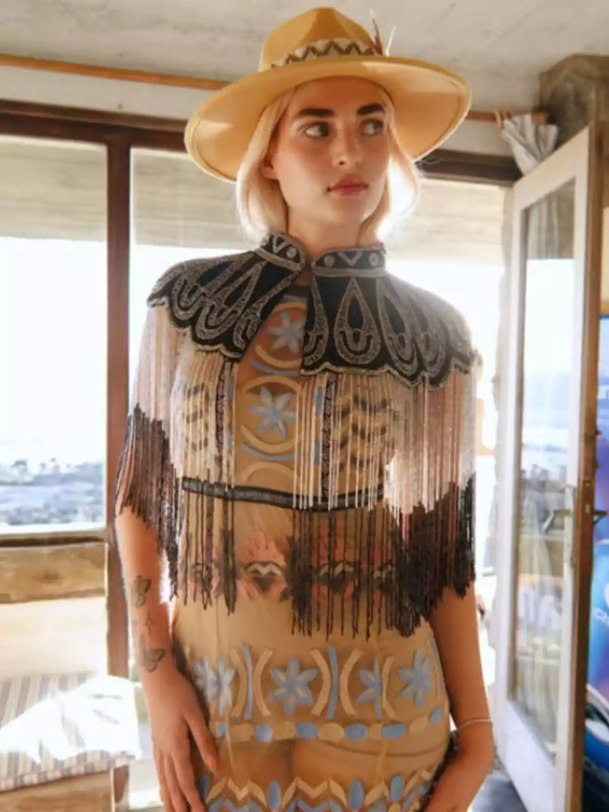 Blonde woman wearing a beige hat and a sheer dress with floral embroidery and a decorative fringed cape in a well-lit indoor setting Blonde woman wearing a beige hat and a sheer dress with floral embroidery and a decorative fringed cape in a well-lit indoor setting