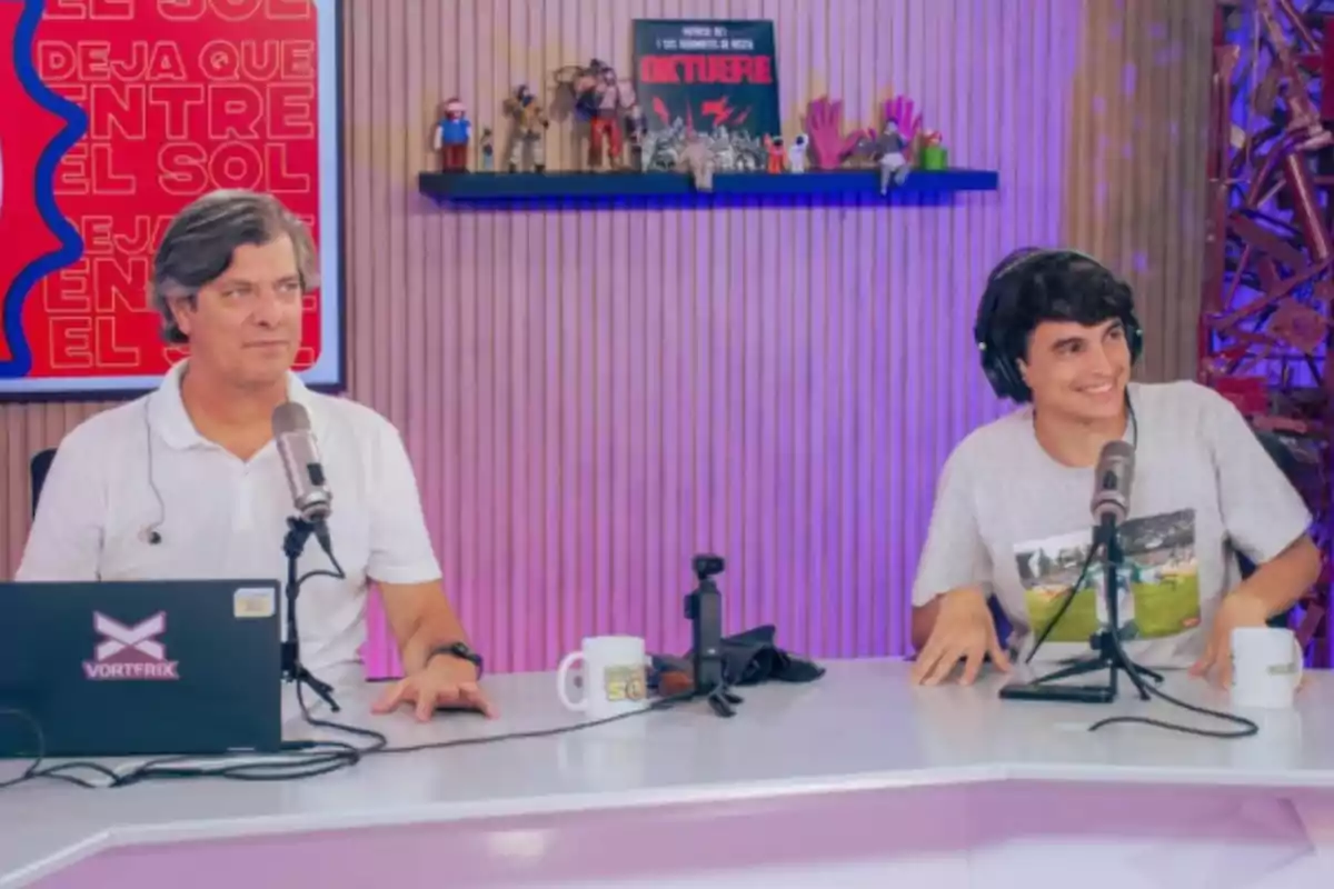 Dos personas sentadas en una mesa de radio con micrófonos y auriculares, una de ellas sonríe mientras la otra mira hacia el frente, al fondo hay una repisa con figuras y un cartel que dice OKTUBRE