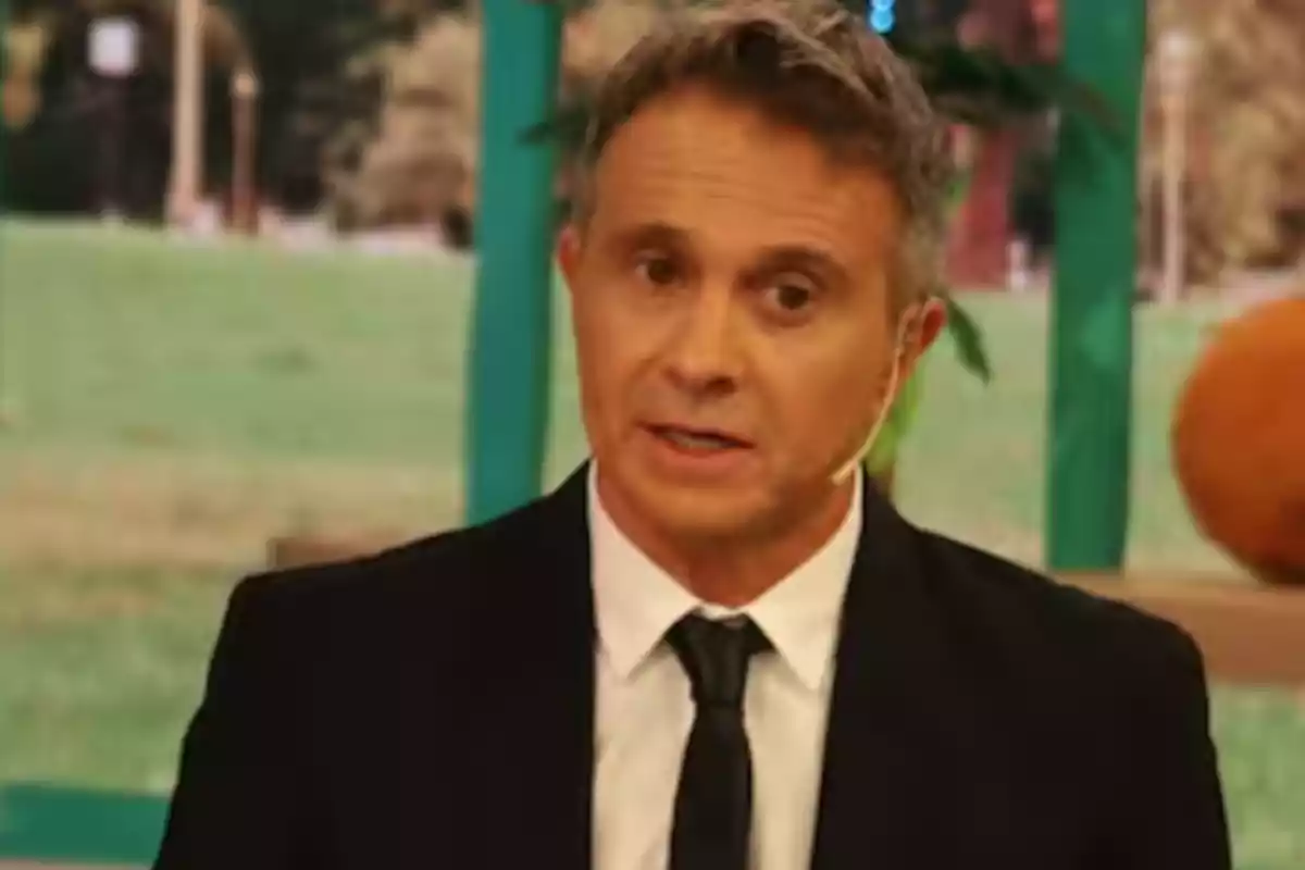 Short-haired, gray-haired man wearing a black suit and tie, speaking on a television set with a blurred green background.