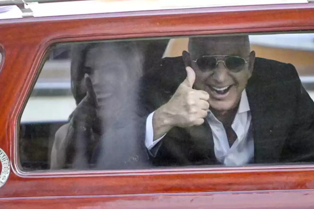 Hombre sonriente con gafas de sol haciendo el gesto de pulgar arriba desde el interior de un vehículo junto a una mujer desenfocada