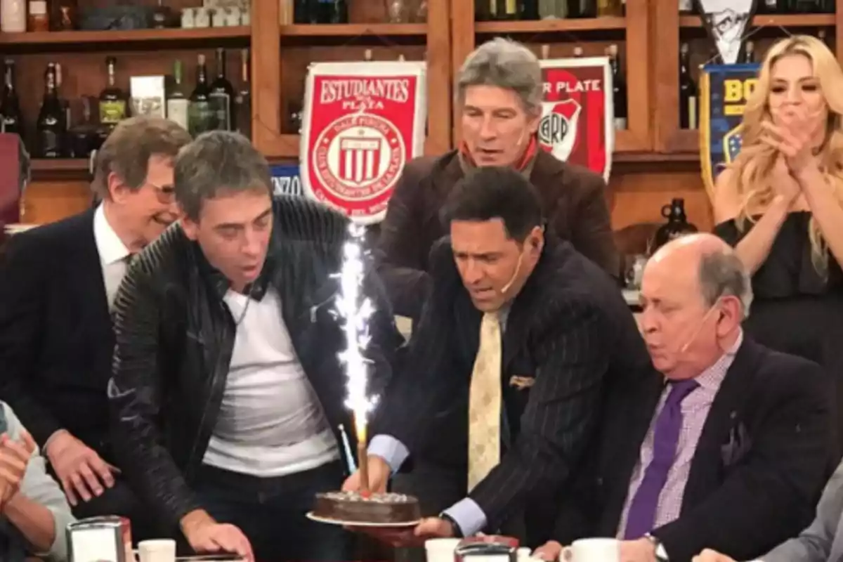 Un grupo de personas celebra un cumpleaños en un estudio de televisión mientras una torta con una vela de bengala está en el centro de la mesa y todos observan con entusiasmo