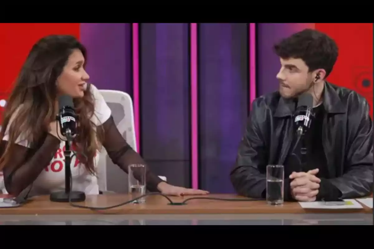 Dos personas conversan frente a micrófonos en un estudio de radio o podcast, sentados en una mesa con vasos de agua. Dos personas conversan frente a micrófonos en un estudio de radio o podcast, sentados en una mesa con vasos de agua.