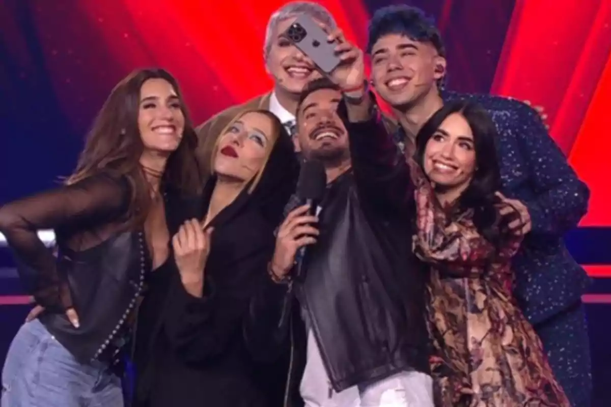 Un grupo de seis personas sonríe mientras se toma una selfie en un escenario iluminado con luces rojas de fondo Un grupo de seis personas sonríe mientras se toma una selfie en un escenario iluminado con luces rojas de fondo
