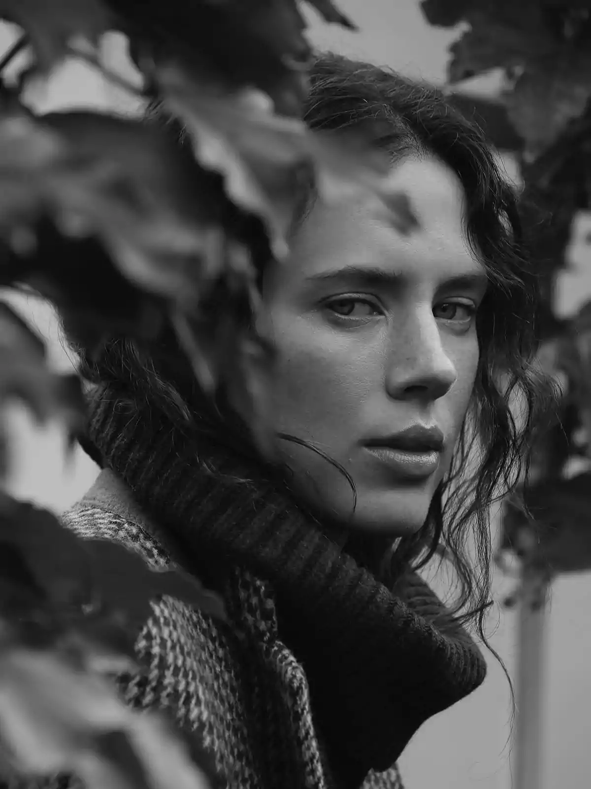Black and white portrait of a person in a turtleneck sweater, surrounded by leaves, looking at the camera with a serious expression Black and white portrait of a person in a turtleneck sweater, surrounded by leaves, looking at the camera with a serious expression
