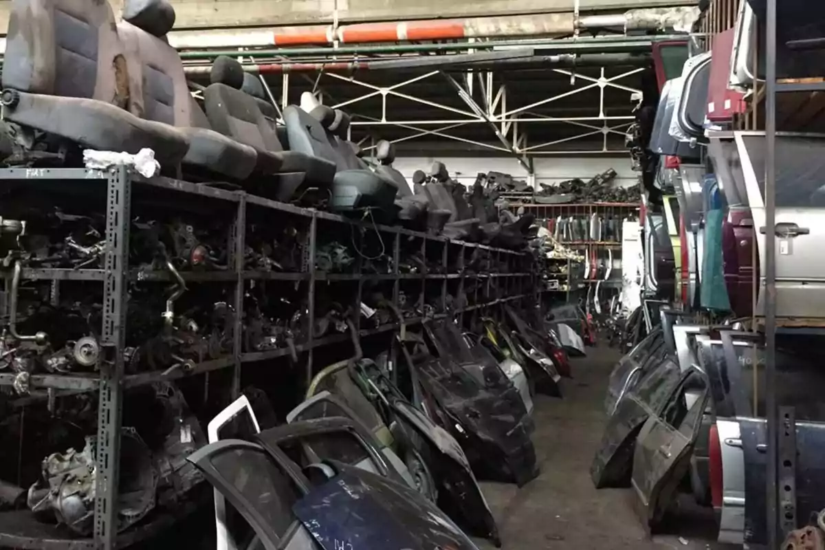 Interior de un deshuesadero de autos con asientos, puertas y piezas de automóviles apiladas en estantes y el suelo
