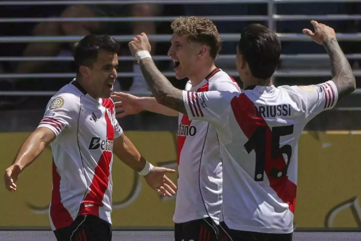 Tres futbolistas con uniforme blanco y una franja roja celebran un gol en un estadio Tres futbolistas con uniforme blanco y una franja roja celebran un gol en un estadio