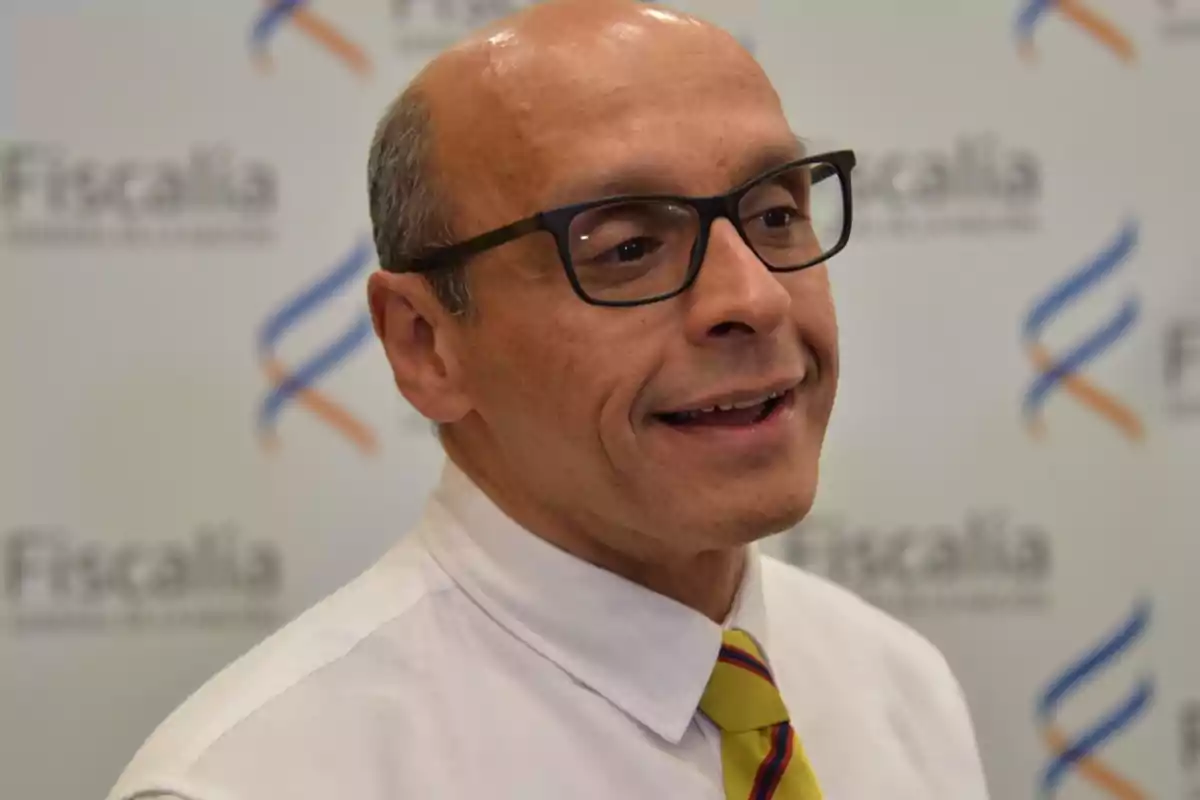 Bald man with glasses and a yellow striped tie, smiling in front of a background with Prosecutor's Office logos Bald man with glasses and a yellow striped tie, smiling in front of a background with Prosecutor's Office logos