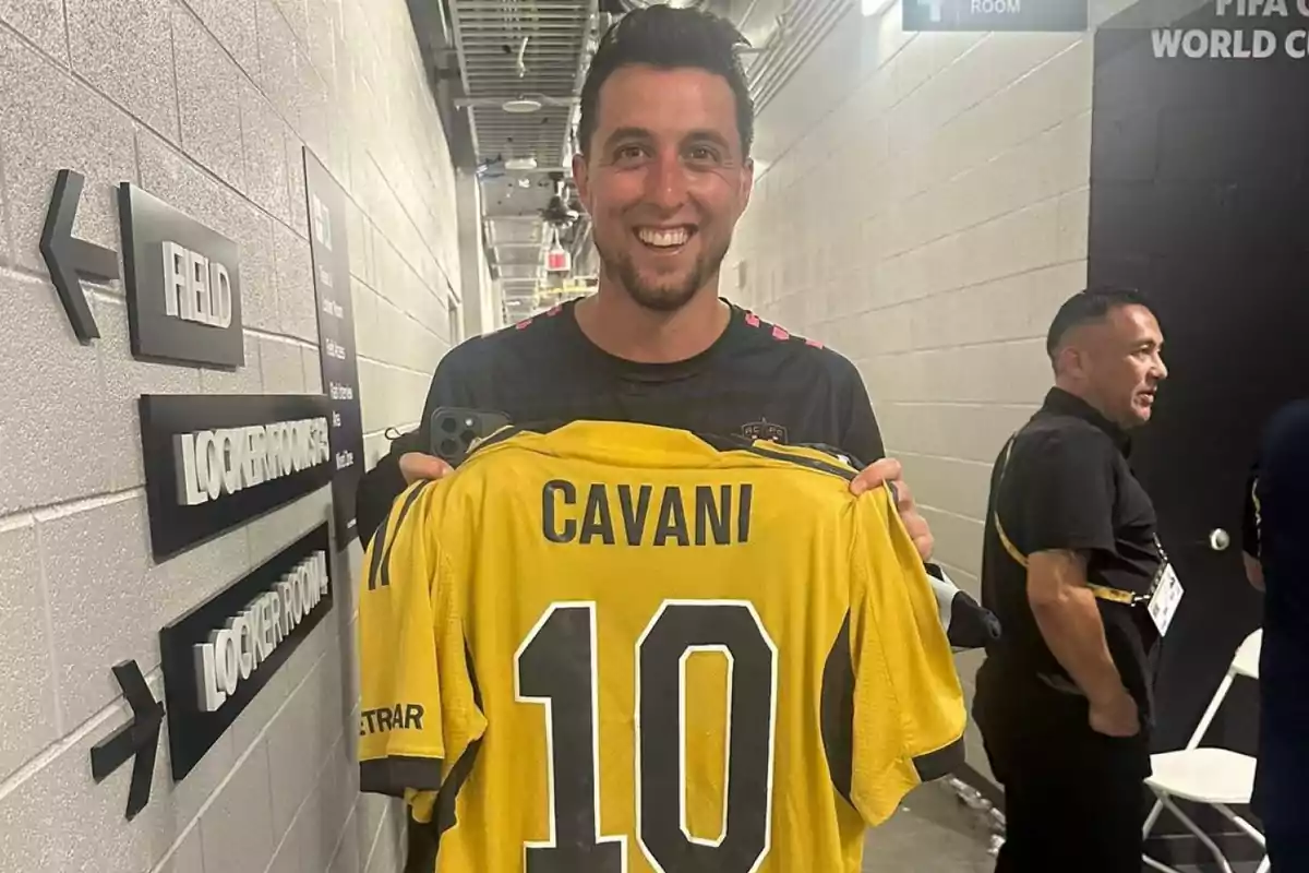 A smiling man holds a yellow soccer jersey with the name Cavani and the number 10 in a stadium hallway. A smiling man holds a yellow soccer jersey with the name Cavani and the number 10 in a stadium hallway.