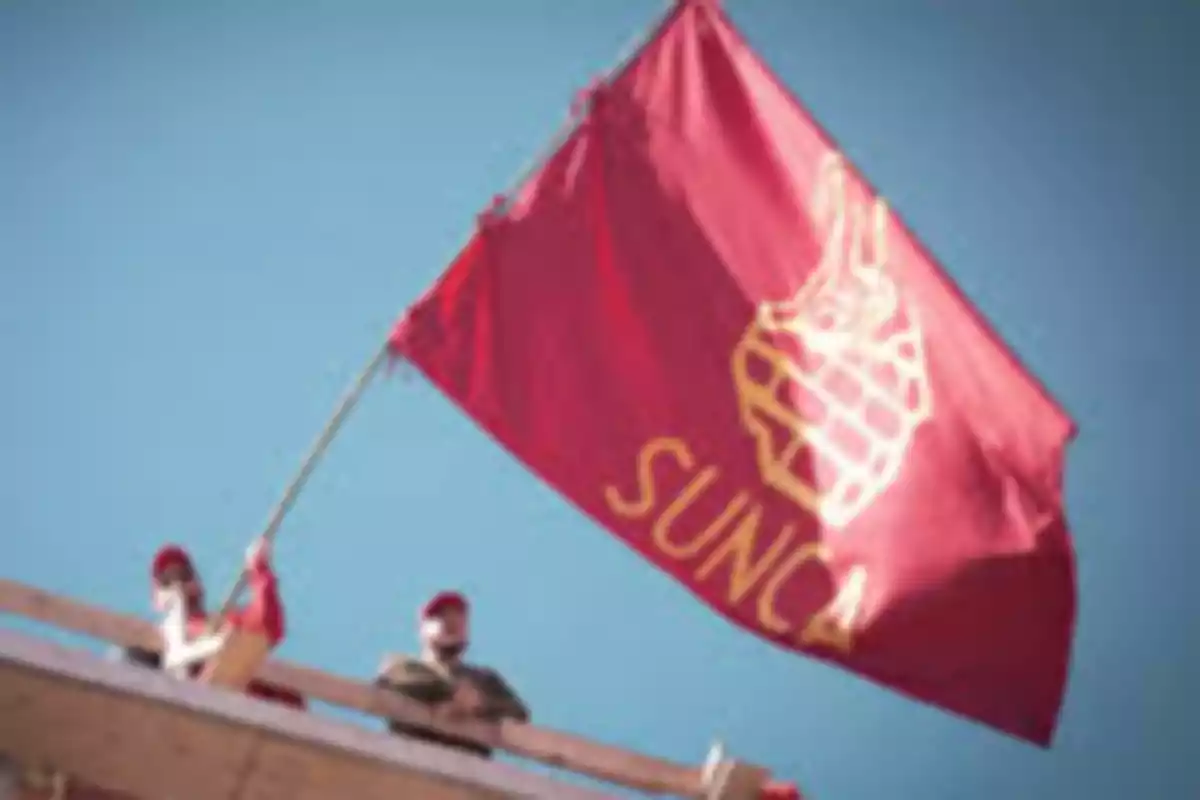 Red flag with the SUNCA logo and name waving atop a building next to two people wearing red caps Red flag with the SUNCA logo and name waving atop a building next to two people wearing red caps