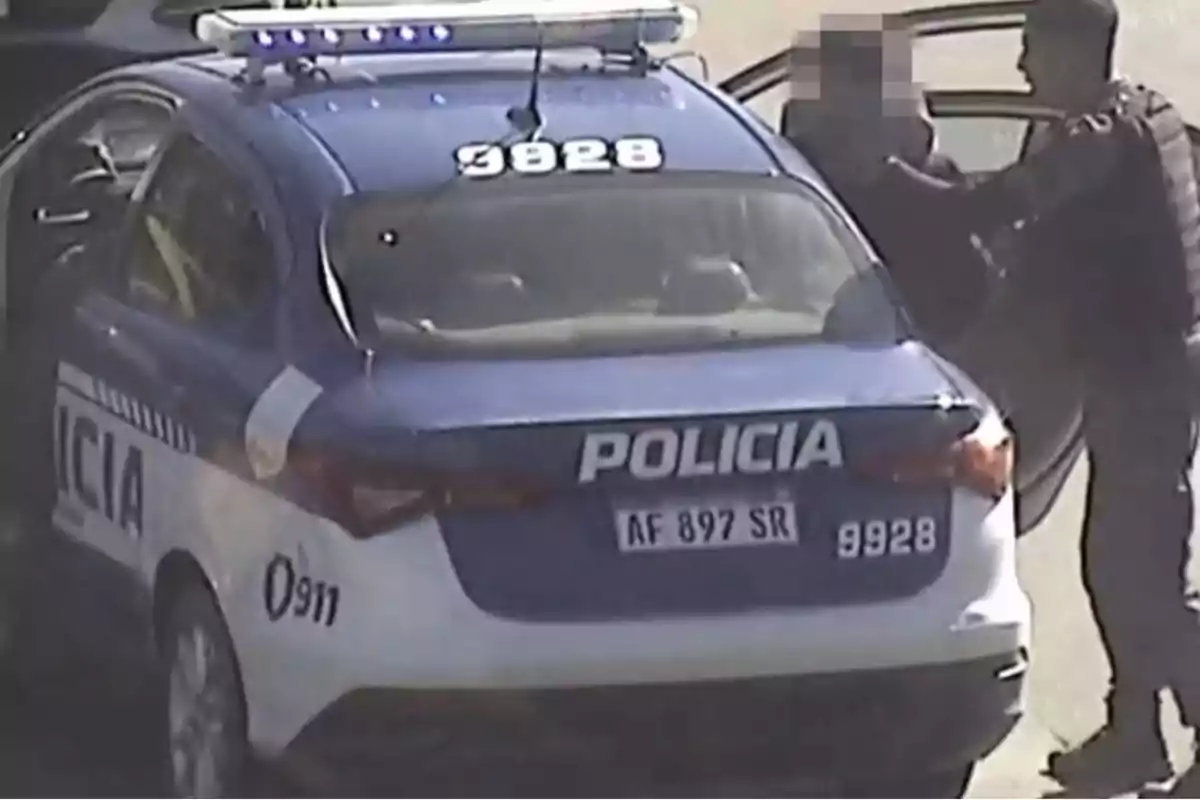 Patrullero de la policía detenido con las puertas abiertas mientras un agente interactúa con una persona junto al vehículo en la vía pública Patrullero de la policía detenido con las puertas abiertas mientras un agente interactúa con una persona junto al vehículo en la vía pública