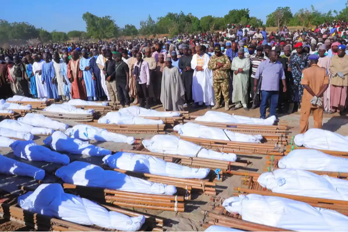 A large group of people gathers outdoors to pay tribute to several bodies wrapped in white cloths and placed on wooden stretchers. A large group of people gathers outdoors to pay tribute to several bodies wrapped in white cloths and placed on wooden stretchers.
