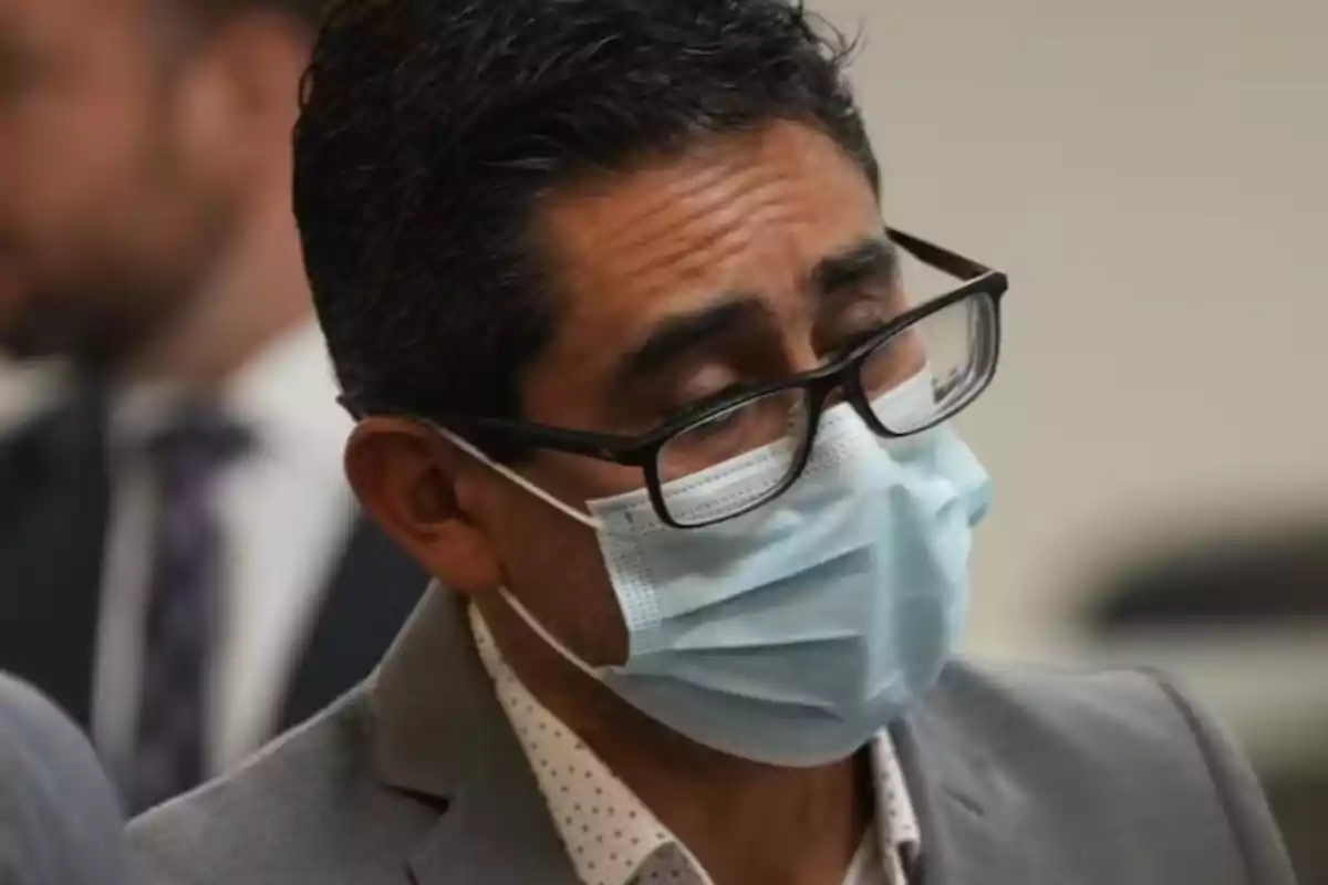 Hombre con gafas y cubrebocas azul viste saco gris y camisa blanca con puntos pequeños Hombre con gafas y cubrebocas azul viste saco gris y camisa blanca con puntos pequeños