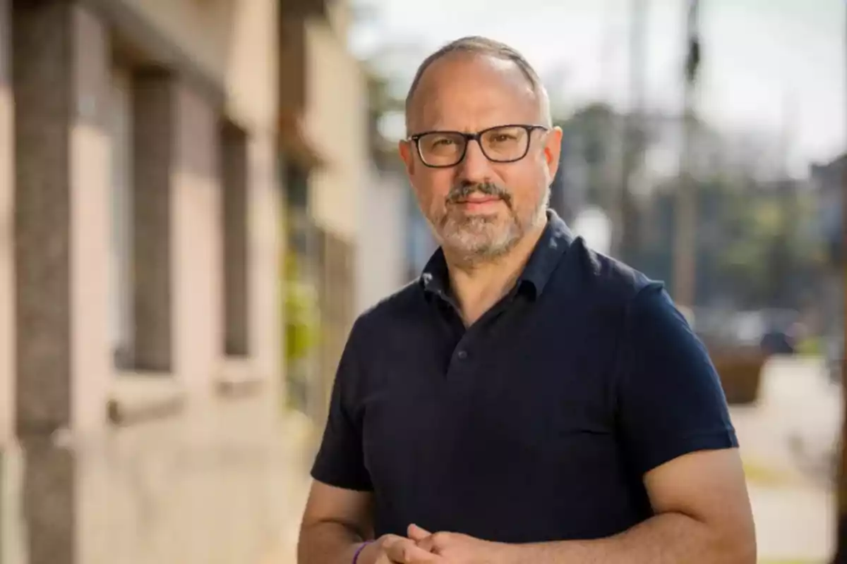 Hombre de mediana edad con gafas y barba, vestido con camiseta oscura, posando al aire libre en una calle urbana durante el día Hombre de mediana edad con gafas y barba, vestido con camiseta oscura, posando al aire libre en una calle urbana durante el día