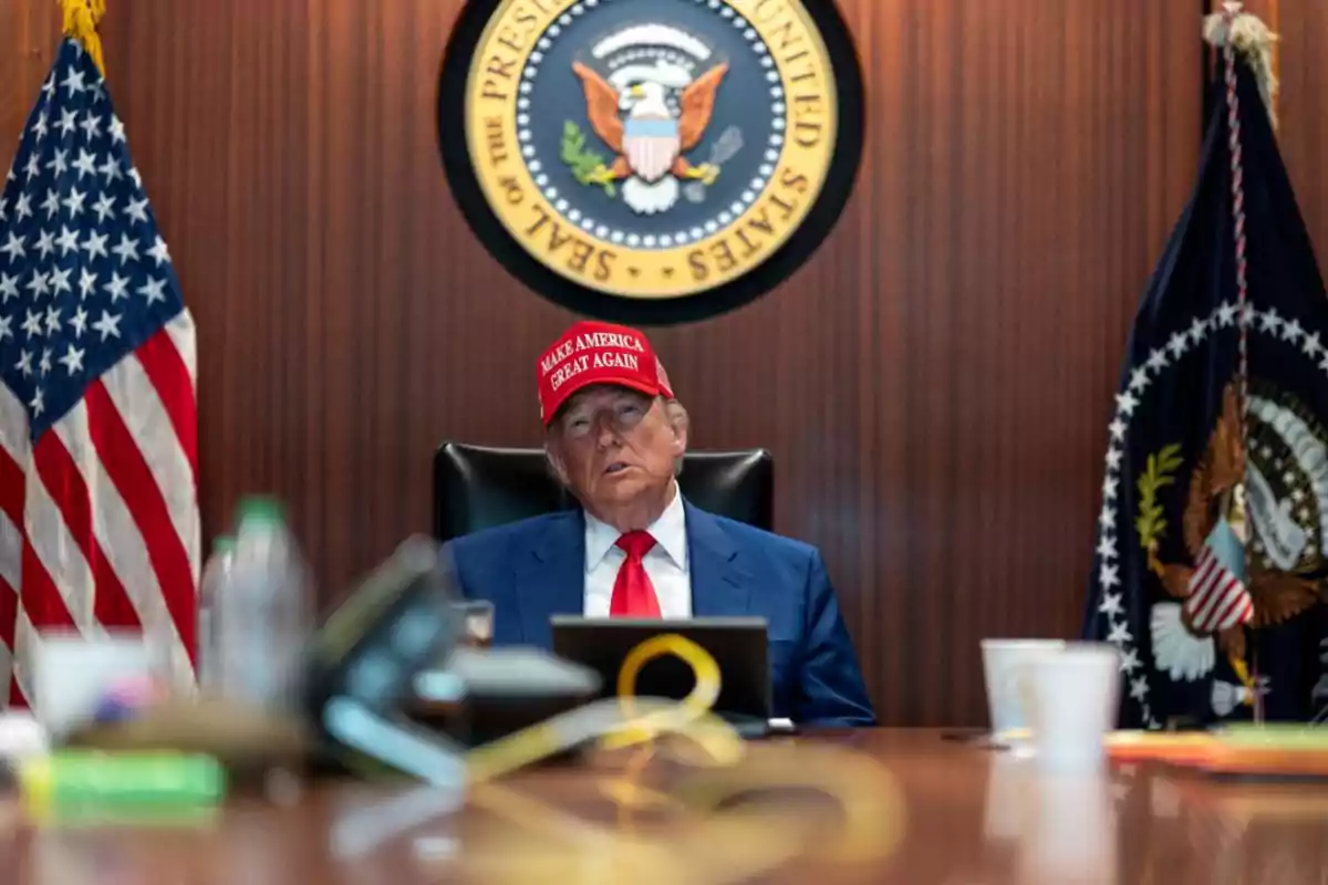 Hombre con gorra roja y traje azul sentado en una sala de reuniones con banderas de Estados Unidos y el sello presidencial al fondo Hombre con gorra roja y traje azul sentado en una sala de reuniones con banderas de Estados Unidos y el sello presidencial al fondo