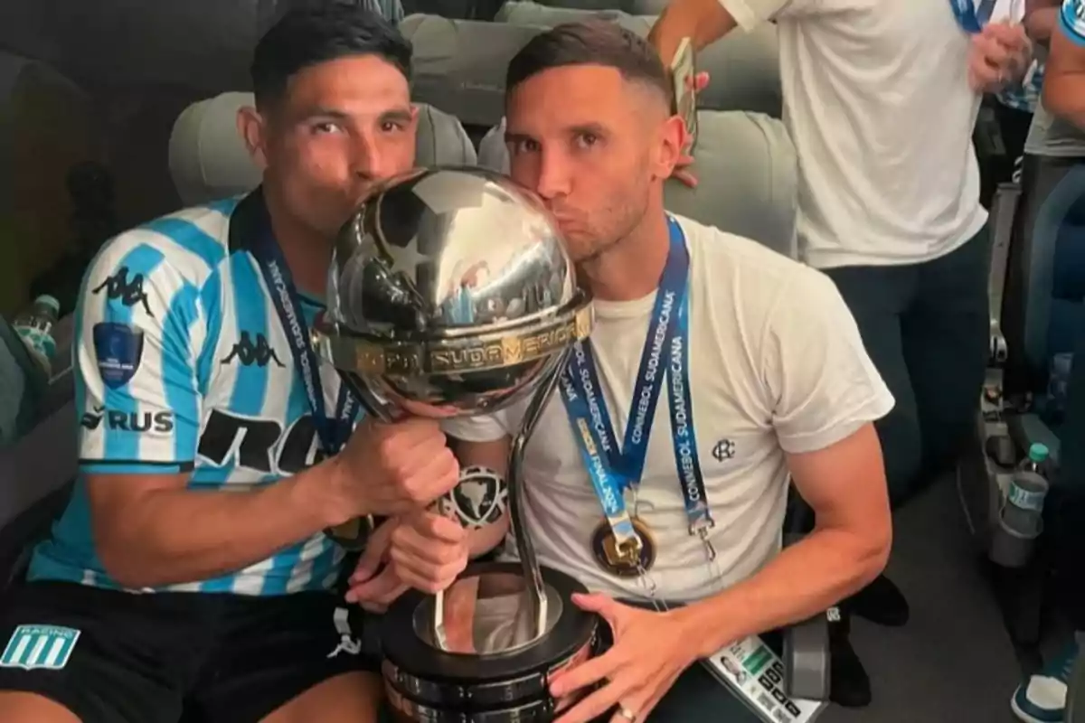 Two players celebrate together by kissing a large silver trophy while sitting on a bus. Two players celebrate together by kissing a large silver trophy while sitting on a bus.