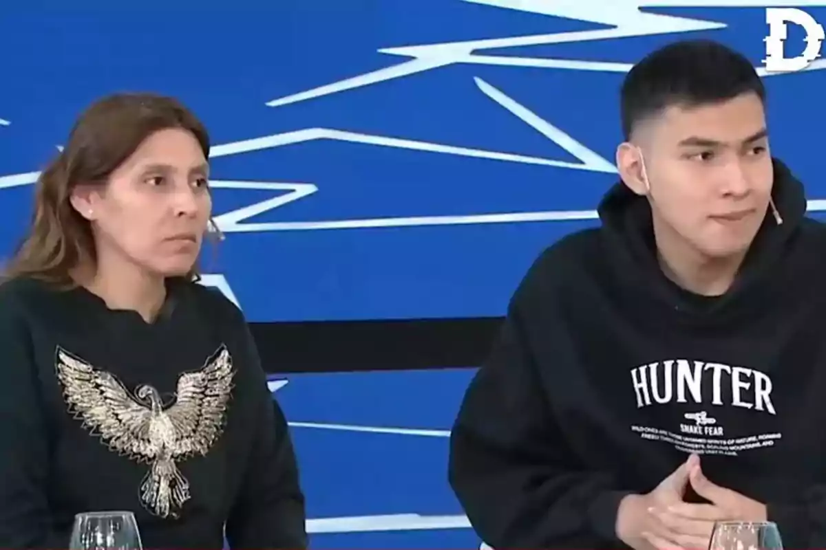 Dos personas sentadas en un set de televisión con fondo azul, una mujer con suéter negro con un águila bordada y un joven con sudadera negra que dice Hunter
