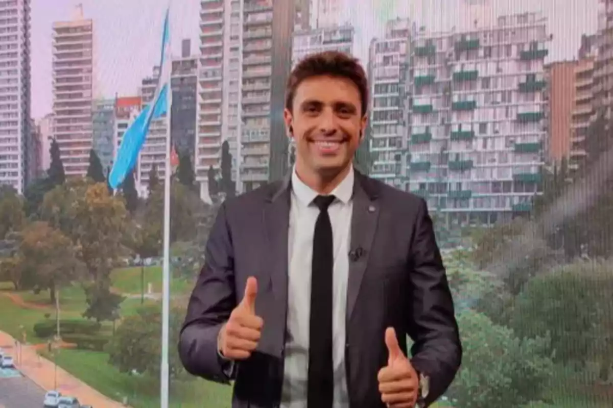 Hombre con traje y corbata sonriendo y haciendo el gesto de pulgares arriba frente a un fondo urbano con edificios y una bandera argentina