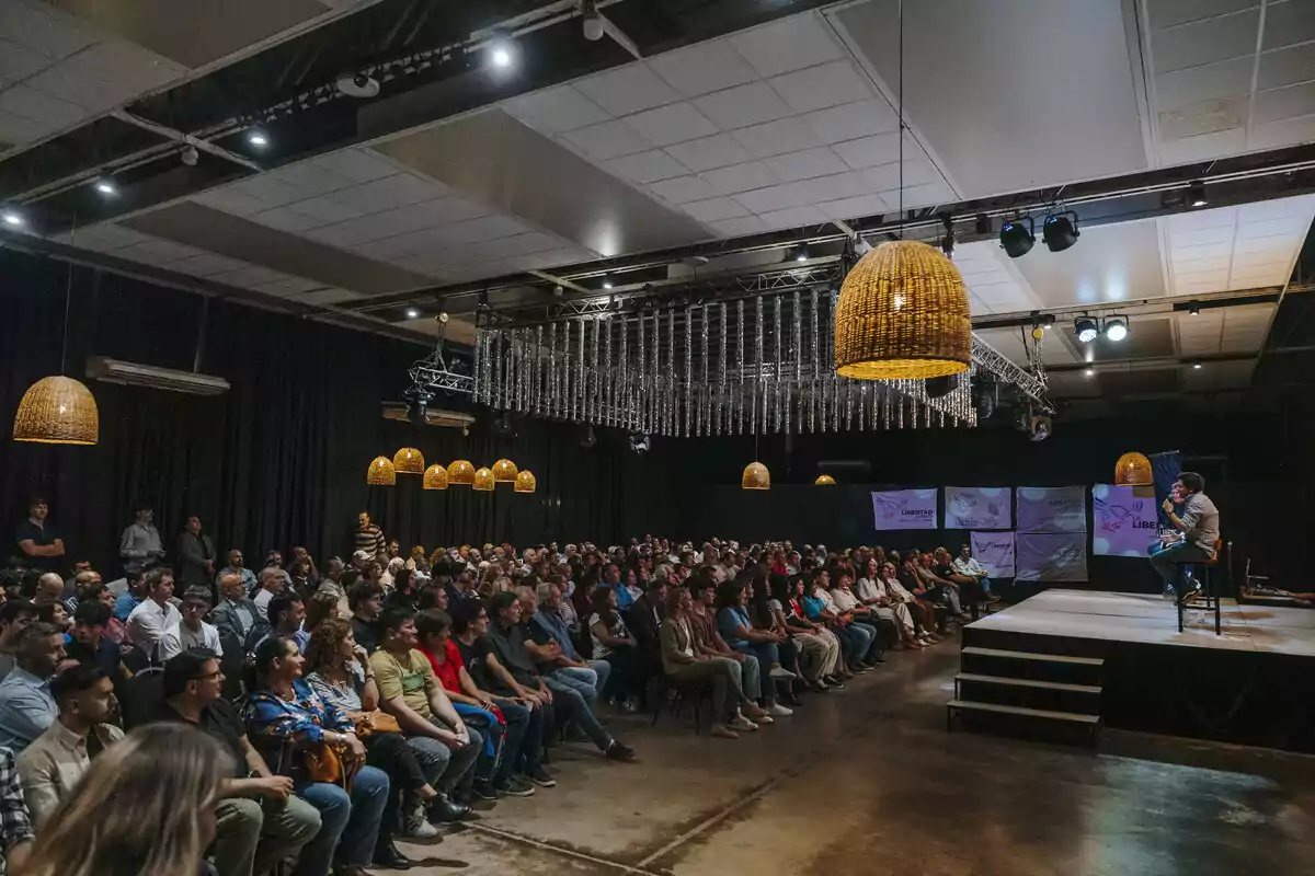 Auditorio lleno de personas sentadas prestando atención a un orador en un escenario iluminado con lámparas colgantes y decoración moderna Auditorio lleno de personas sentadas prestando atención a un orador en un escenario iluminado con lámparas colgantes y decoración moderna